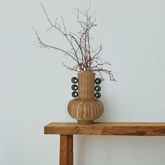 El yapımı rattan vazo, her iki yanında siyah dekoratif halkalar bulunan heykelsi formda; ahşap bank üzerinde, kurumuş dallarla dekore edilmiş