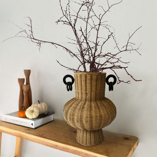 İki kulplu el yapımı rattan vazo; kurumuş dallarla dekore edilmiş, ahşap sehpa üzerinde, yanında ahşap objeler ve dekoratif kitaplarla birlikte